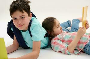 Caucasian smiling handsome teen schoolboy with backpack and workbooks, lying near his younger sister on white background photo