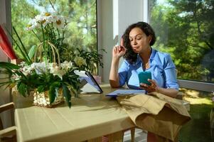 Thoughtful perplexed woman florist, entrepreneur holding mobile phone, working in her workshop in a floral design studio photo