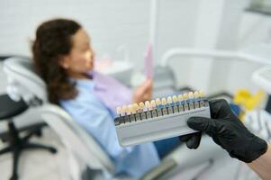 Vita scale. Bleaching teeth guide. Tooth color chart in dentist's hands over blurred patient in dental chair. Dentistry photo