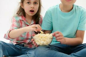 Cropped view kids with a bowl of popcorn, looking fascinated while watching movie, isolated on white studio background photo