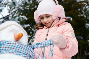 Cheerful lovely child girl in pink down jacket and fluffy earmuffs, putting on a blue scarf around the snowman's neck photo