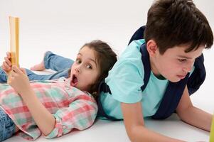 Preschooler girl or first grader with WOW emotion, looking at camera, lying next to her brother with a workbook in hands photo