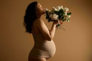 hermosa grávido mujer con grande barriga en el embarazo 28 semana, vistiendo beige lencería, posando con blanco floreciente lilas foto