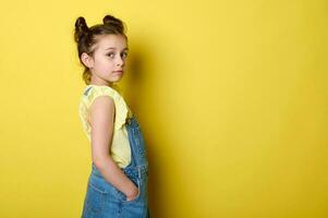 Lovely primary school student girl with stylish looking at camera, standing with hands in pockets over yellow background photo