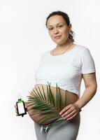 Gorgeous multi ethnic pregnant woman holding exotic palm leaf cosmetic oil massage over her big belly, isolated on white photo