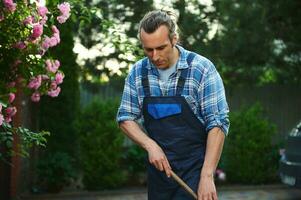 cintura arriba retrato de un hermoso caucásico hombre jardinero en trabajo uniforme, limpieza el malas hierbas desde el losas en patio interior foto