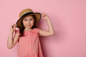 Beautiful little child girl wearing stylish pink dress and straw hat, smiles looking at camera, isolated pink background photo