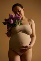 Studio portrait of a beautiful adult pregnant woman caressing her belly, holding a bunch of lilacs, isolated on beige photo