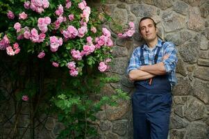 sonriente contento hermoso hombre, jardinero florista en azul trabajo uniforme, mirando a cámara, en pie en un la mansión patio interior foto