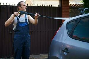 Young adult man directing hose with spray wand on the roof of his car, washing vehicle using water high pressure washer photo