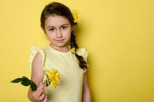 Caucasian lovely baby girl holding a yellow rose flower, looking at camera, isolated on yellow background. Copy ad space photo