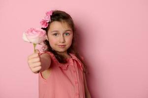 hermosa pequeño niño niña en elegante vestido, participación fuera cámara un hermosa Rosa flor, sonriente terminado rosado antecedentes foto