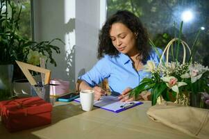 Beautiful woman writing on clipboard, working in cozy small floral shop. Flowwer arrangements and gifts concept photo