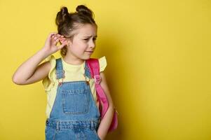 Surprised little girl with backpack, holding hand to her ear, absorbing gossips, expressing bewilderment yellow backdrop photo