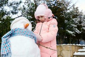 encantador pequeño niño niña pone en un azul chal en un muñeco de nieve, durante activo invierno ocio juegos en el Nevado patio interior foto