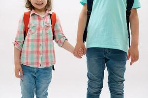 Close-up teen boy and preschooler girl holding hands, isolated on white backdrop. Kids with backpack going to the school photo