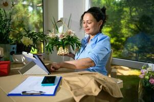 Pleasant female florist holding a wicker basket with flower arrangement, talking with clients via video link on tablet photo