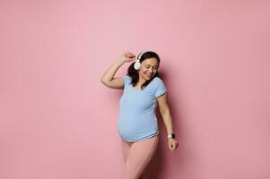 Cheery pregnant woman dancing listening to music on headphones, enjoying wonderful moments of pregnancy isolated on pink photo
