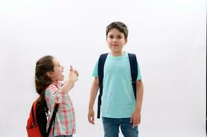 Smart school kids with backpacks, mischievous little girl smiles looking at her older brother, isolated white background photo