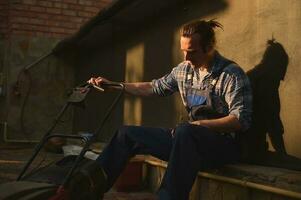 Portrait of an overworked gardener in blue overalls with a lawn mower in the garden at sunset after a hard day's work photo