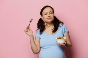 Delightful expectant pregnant woman eating fruits salad, isolated pink background. Healthy nutrition in pregnancy time photo