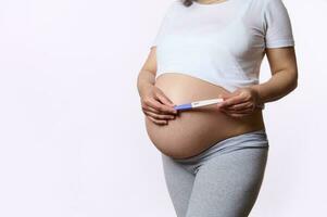 Close-up gravid woman, expectant mother holding pregnancy inkjet test above her pregnant belly, isolated white backdrop photo