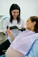 Pregnant woman patient at dental appointment, sitting in dentist's chair, smiling in mirror, discussing with the doctor photo