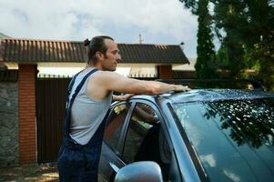 Young attractive Latin American man in blue overalls, manually washing his car outdoors with cleaning foam and sponge. photo