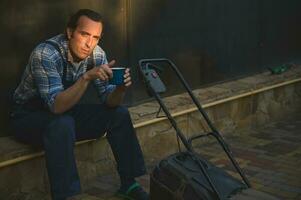 Portrait of a gardener man looking at camera, sitting near a lawn mower with cup of coffee in hands, relaxing at sunset photo