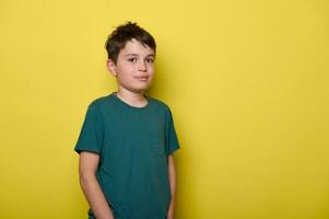 Handsome male teenager boy smiles looking at camera, standing on isolated yellow background with his hands in pockets photo