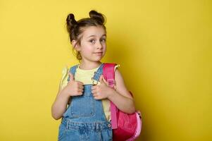 encantador pequeño niña con mochila, pulgar arriba a cámara, alegría a el comienzo de el colegio año en el primero grado foto