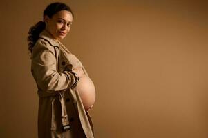 Beautiful pregnant woman in beige trench coat, caressing her big bare belly in late pregnancy, looking at camera photo