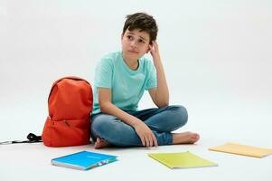 Authentic teen school kid sitting near orange backpack and textbooks, reasoning over school problems, isolated on white photo