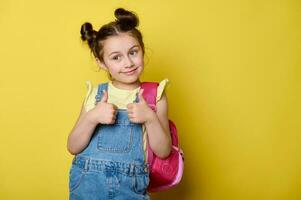 Cute child girl in yellow t-shirt and blue denim dress overalls, gesturing thumbs up, smiling over yellow background photo