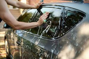 Close-up hands of a car service worker rubbing soapy detergent foam into passenger rear window while washing car by hand photo