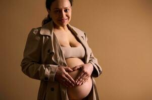 hermosa embarazada mujer en beige zanja abrigo, formando un corazón forma con manos terminado su desnudo barriga en beige antecedentes foto