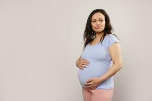 Perplexed pregnant woman expecting baby, holding hands on belly, thoughtfully looking aside, isolated white background photo