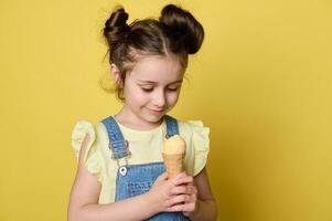 contento pequeño niña disfrutando dulce verano postre, participación gofre cono con hielo crema cucharón, aislado en amarillo antecedentes foto