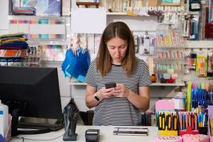Young woman manager, seller in creative store, using mobile phone, standing at the counter and ordering stationery photo