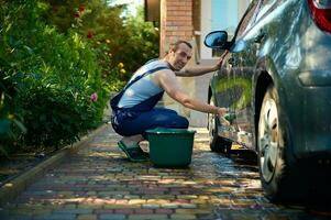 Handsome young adult man in blue coverall washing his car with foamy detergent and sponge rag, smiling looking at camera photo