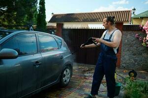 Handsome muscular attractive young adult man in blue work overalls, manually washing car under high pressure water jet photo