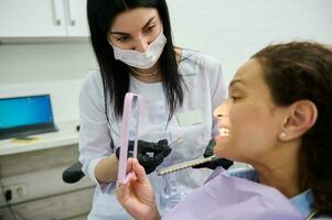 hembra paciente sentado en del dentista silla, sonrisas mirando a espejo mientras visitando dentista para dientes blanqueamiento procedimiento foto