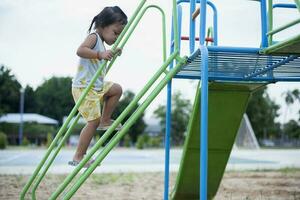 un niña es alpinismo arriba un acero escalera a un diapositiva en un patio de juegos. foto