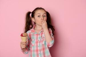 Emotional little girl with ponytails, holding a waffle cone with ice cream, kissing finger, showing delicious hand sign photo