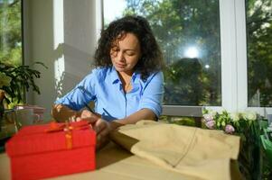 Beautiful curly haired Latin American woman taking notes in a floral workshop. Beige apron and red gift box on the table photo