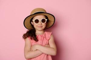 elegante de moda pequeño niña en Gafas de sol, rosado vestir y Paja sombrero, posando con brazos doblada en rosado antecedentes foto