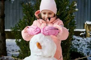 sonriente pequeño niña jugando al aire libre invierno juegos, edificio un grande monigote de nieve en el patio interior, vistiendo calentar rosado abajo chaqueta foto