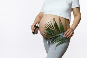 Close-up gravid woman holding palm leaf above her pregnant belly, applying massage oil to reduce prevent stretch marks photo