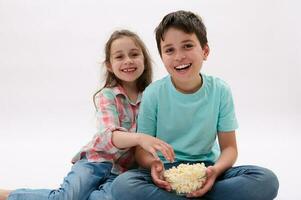 Caucasian happy children sitting over white isolated studio background, watching cartoons or movie, eating popcorn. photo