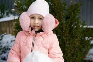 hermosa pequeño niña en rosado mullido oído manguitos y abajo chaqueta, sonriente cámara, en pie cerca un monigote de nieve en el patio interior foto